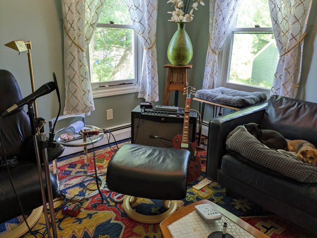 A Fender amp, Les Paul Studio, and drum machine sit in the corner of a room, framed by two windows. Two dogs sleep on a nearby couch.